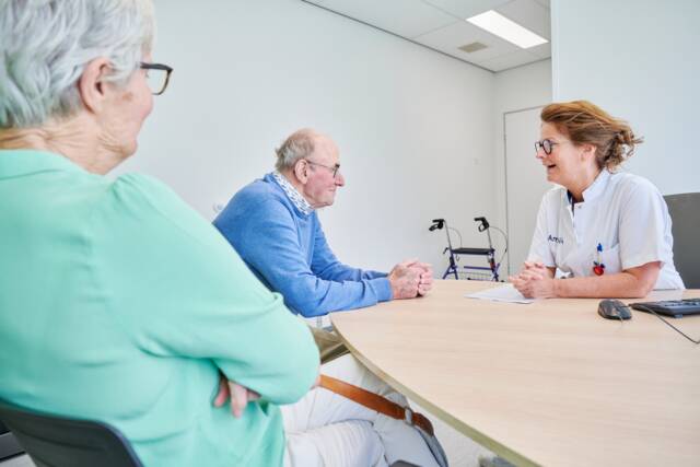 zorgverlener praat met man aan tafel, vrouw zit ernaast
